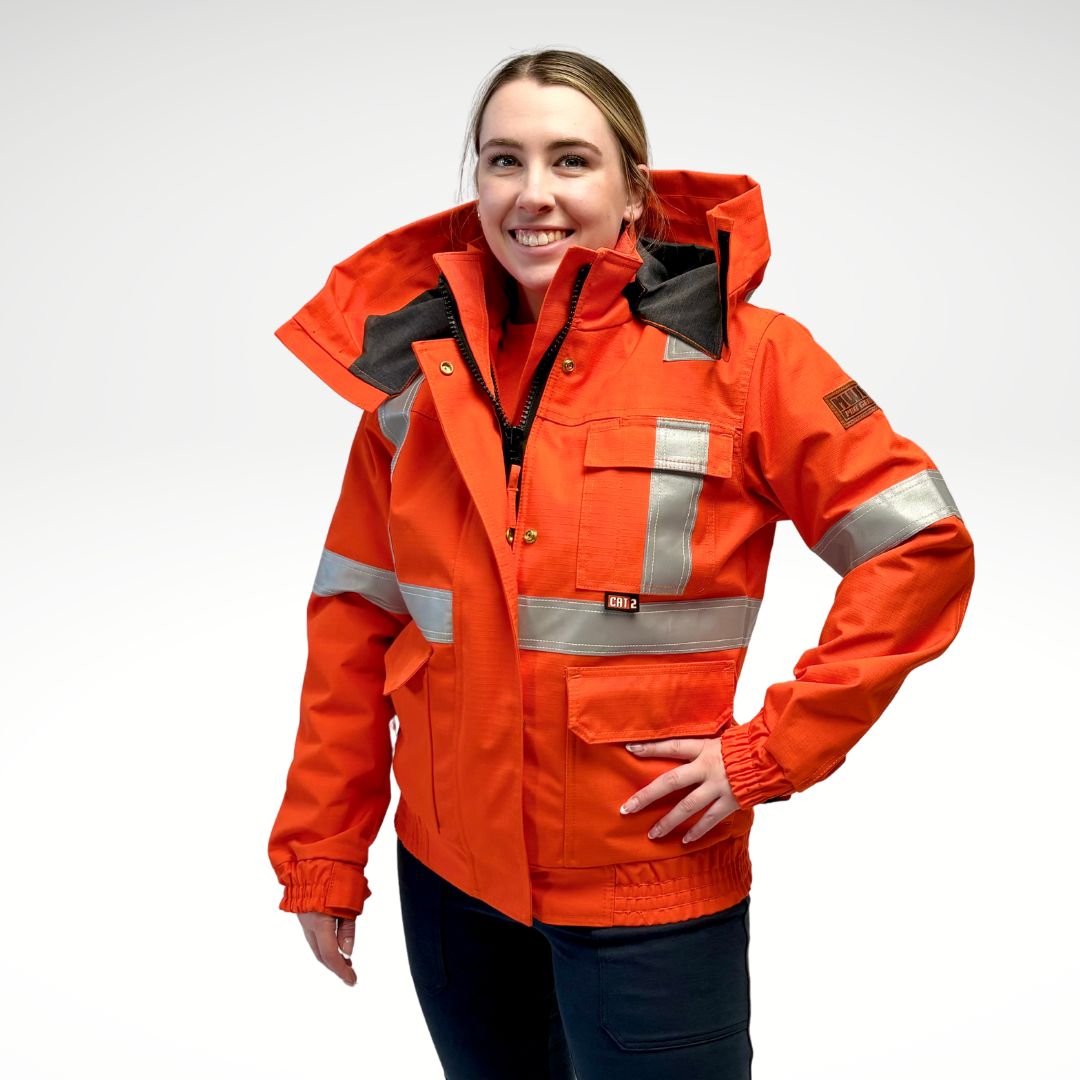 Image of women's MWG FR Bomber Jacket. Women's FR Bomber Jacket is bright orange in colour with silver reflective striping on arms and torso to meet high-visibility standard CSA Z96-15. Image displays elastic cuffs and waistband, 3 large front pockets, detachable hood, and grey hood lining. Women's FR Jacket is made with an inherent flame-resistant (FR) fabric. Women's FR freezer jacket zips in for additional warmth.