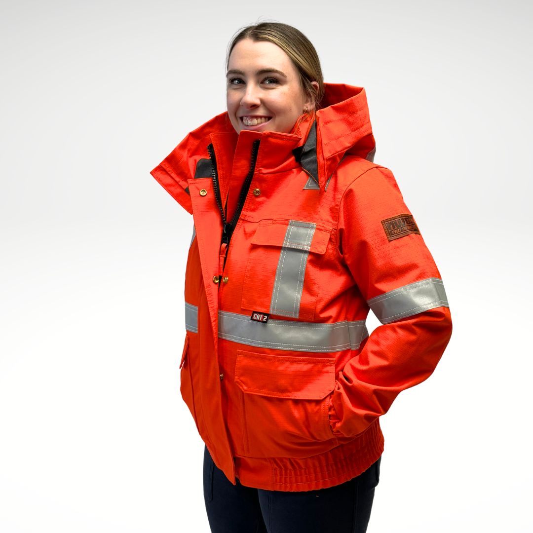 Image of women's MWG FR Bomber Jacket. Women's FR Bomber Jacket is bright orange in colour with silver reflective striping on arms and torso to meet high-visibility standard CSA Z96-15. Image displays elastic cuffs and waistband, 3 large front pockets, detachable hood, and grey hood lining. Women's FR Jacket is made with an inherent flame-resistant (FR) fabric. Women's FR freezer jacket zips in for additional warmth.