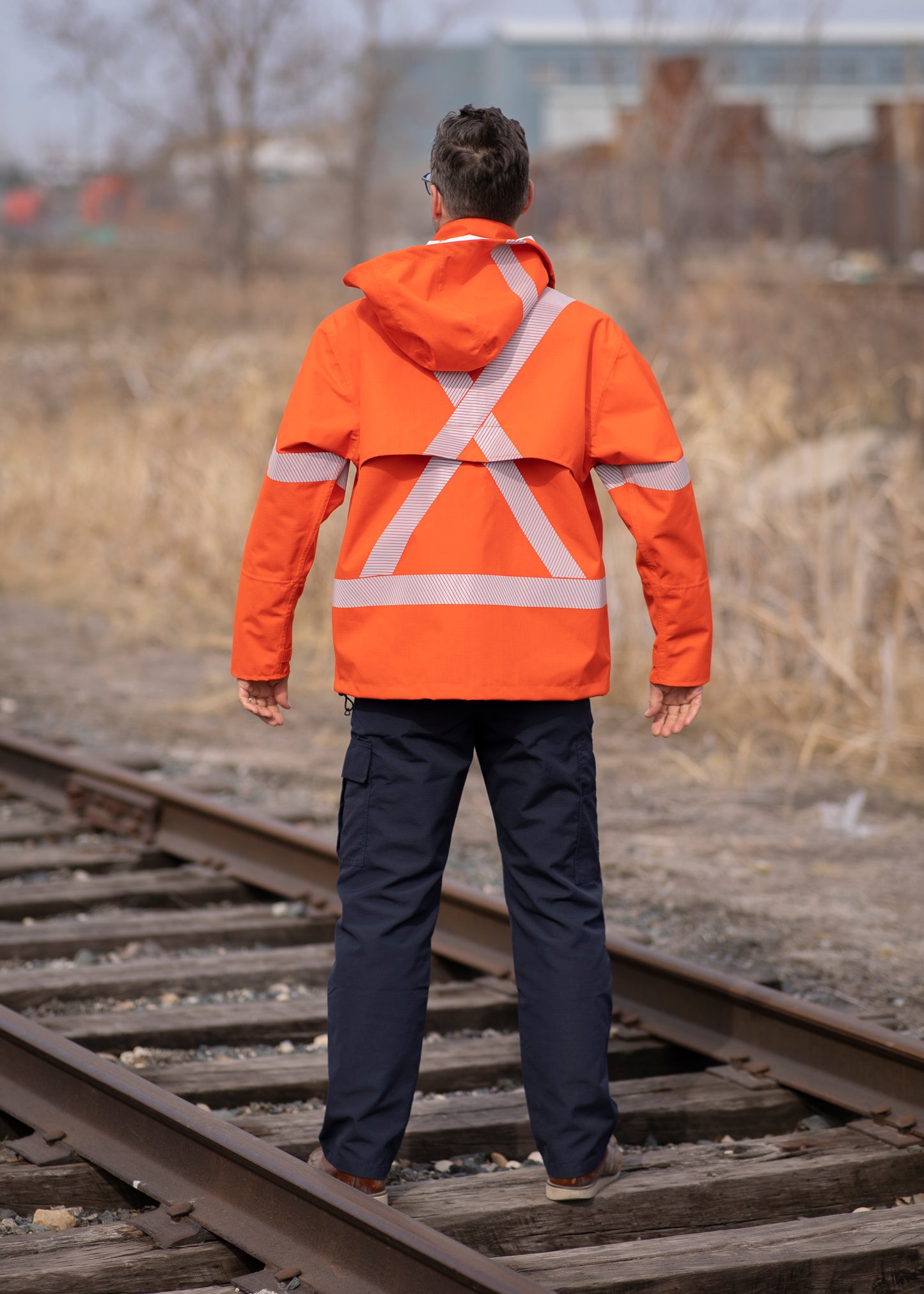 Back view of MWG STORMSHIELD FR Rain Jacket. Image displays the vented back and silver segmented reflective tape in an X pattern to meet high-visibility standard CSA Z96-15. Image shows the detachable hood hanging on the back of the model. Model is wearing MWG STORMSHIELD FR Rain Jacket with navy MWG RIPGUARD FR Utility Pant.