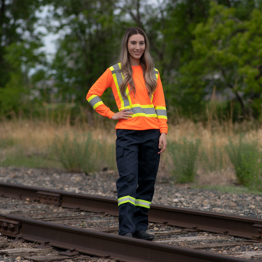 Image of MWG Women's Hi-Vis Long Sleeve T-Shirt. Women's Long Sleeve shirt is bright orange with yellow/silver/yellow reflective tape on torso and sleeves to meet high-visibility standard CSA Z96-15. Model is wearing Hi-Vis Shirt with navy Hi-Vis cargo pants.