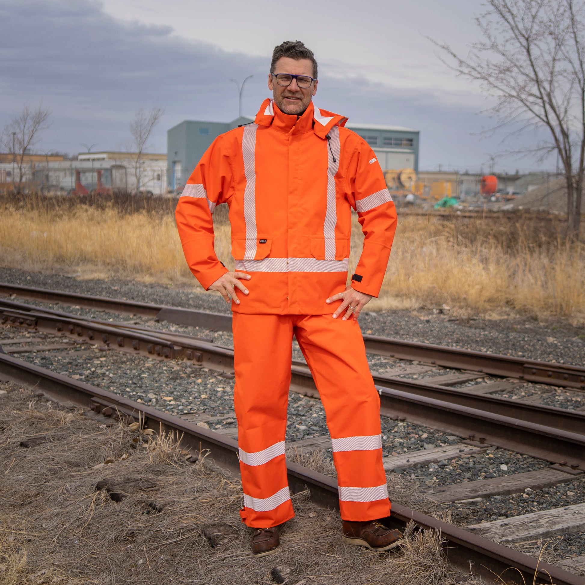 Image of MWG STORMSHIELD FR Rain Proof Jacket. MWG STORMSHIELD FR Rain Proof Jacket is bright orange in colour with silver segmented reflective tape on torso and arms to meet high-visibility standard CSA Z96-15. MWG STORMSHIELD FR Rain Jacket has two front pockets, a detachable hood, and velcro closures on cuffs. Model is wearing MWG STOMSHIELD FR rain jacket with orange MWG STORMSHIELD FR rain proof bib.