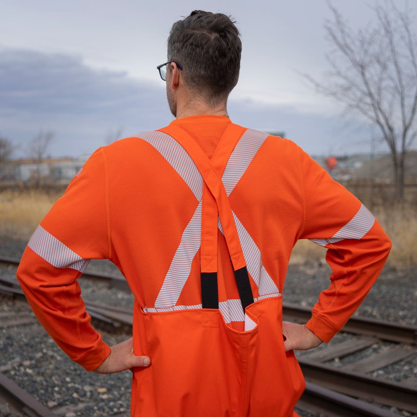 Back view of MWG STORMSHIELD FR Rain Proof Bib. Image displays the orange back straps on the bib. Model is wearing the STORMSHIELD FR Bib over MWG EVOLUTION FR long-sleeve t-shirt. MWG STORMSHIELD is a laminated inherent flame-resistant (FR) fabric.