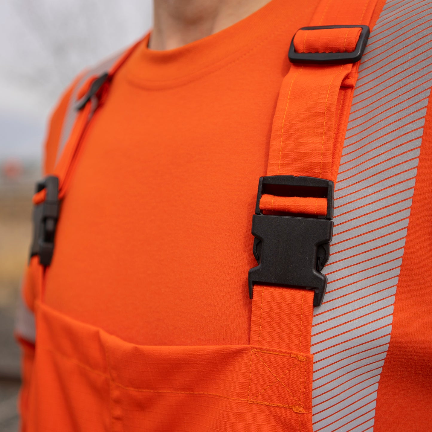 Close up image of the front shoulder straps on MWG STORMSHIELD FR Rain Proof Bib. Front straps include black melamine buckles for adjustability. Model is wearing MWG STORMSHIELD FR Bib over bright orange MWG EVOLUTION FR long-sleeve shirt. MWG STORMSHIELD is a laminated inherent flame-resistant (FR) fabric.