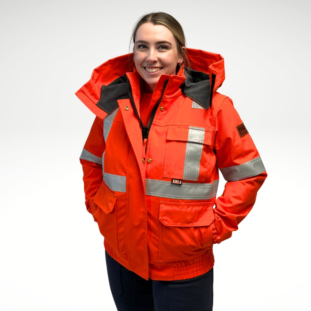 Image of women's MWG FR Bomber Jacket. Women's FR Bomber Jacket is bright orange in colour with silver reflective striping on arms and torso to meet high-visibility standard CSA Z96-15. Image displays elastic cuffs and waistband, 3 large front pockets, detachable hood, and grey hood lining. Women's FR Jacket is made with an inherent flame-resistant (FR) fabric. Women's FR freezer jacket zips in for additional warmth.