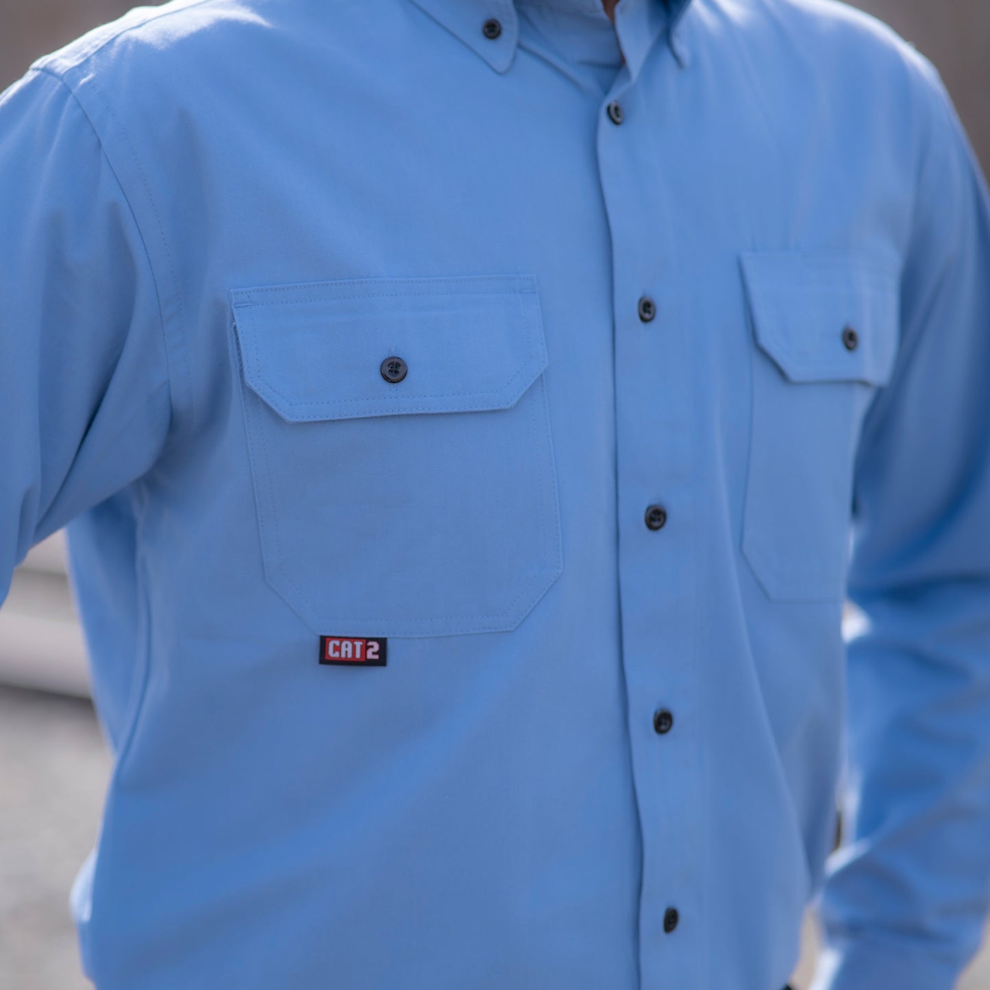 Close up image of MWG COMFORT WEAVE Uniform Shirt in light blue. MWG FR shirt has black buttons and two large chest pockets with flap closures. Image displays CAT 2 identification, symbolizing level of flame-resistance.