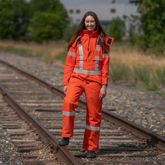 Image of MWG women's FR bib overall. Women's FR Overalls are bright orange in colour with silver reflective tape on torso and black shoulder straps. Model is wearing overalls on top of women's MWG BLOCKER full-zip hoodie. Women's FR Overalls are made with an inherent flame-resistant (FR) fabric.