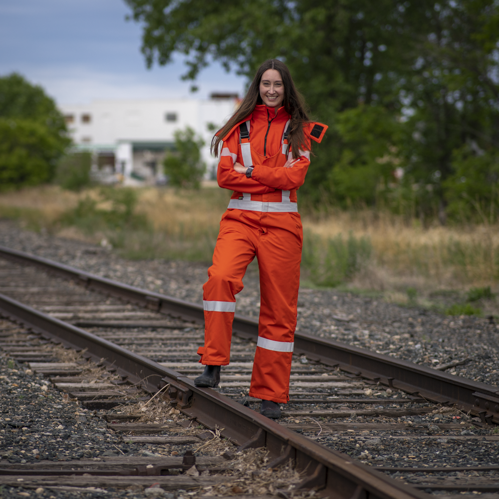 Image of MWG women's FR bib overall. Women's FR Overalls are bright orange in colour with silver reflective tape on torso and black shoulder straps. Model is wearing overalls on top of women's MWG BLOCKER full-zip hoodie. Women's FR Overalls are made with an inherent flame-resistant (FR) fabric.