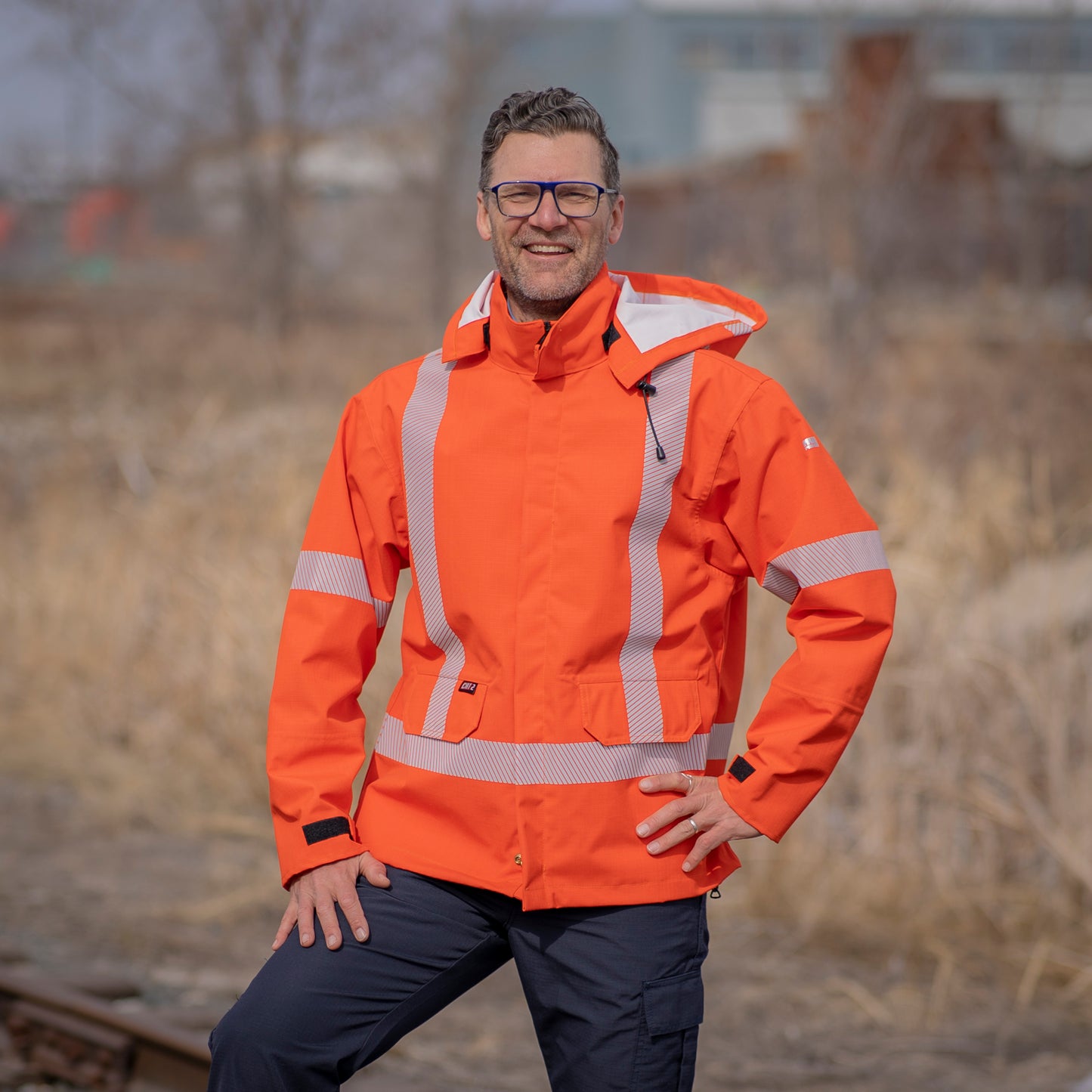 Image of MWG STORMSHIELD FR Rain Jacket. FR Rain Jacket is bright orange with silver segmented reflective tape on torso and sleeves to meet CSA Z96-15 high-visibility standard. MWG STORMSHIELD is an inherent flame-resistant (FR) fabric that's been laminated for wind and waterproofness. Model is wearing FR Rain Jacket with navy MWG RIPGUARD FR Pants.