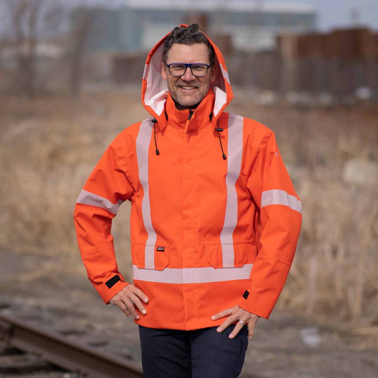 Image of MWG STORMSHIELD FR Rain Proof Jacket. MWG STORMSHIELD FR Rain Proof Jacket is bright orange in colour with silver segmented reflective tape on torso and arms to meet high-visibility standard CSA Z96-15. Model is wearing MWG FR Rain Jacket with hood up, displaying the black drawstrings and white inner lining. MWG STORMSHIELD is a lightweight laminated flame-resistant (FR) fabric.