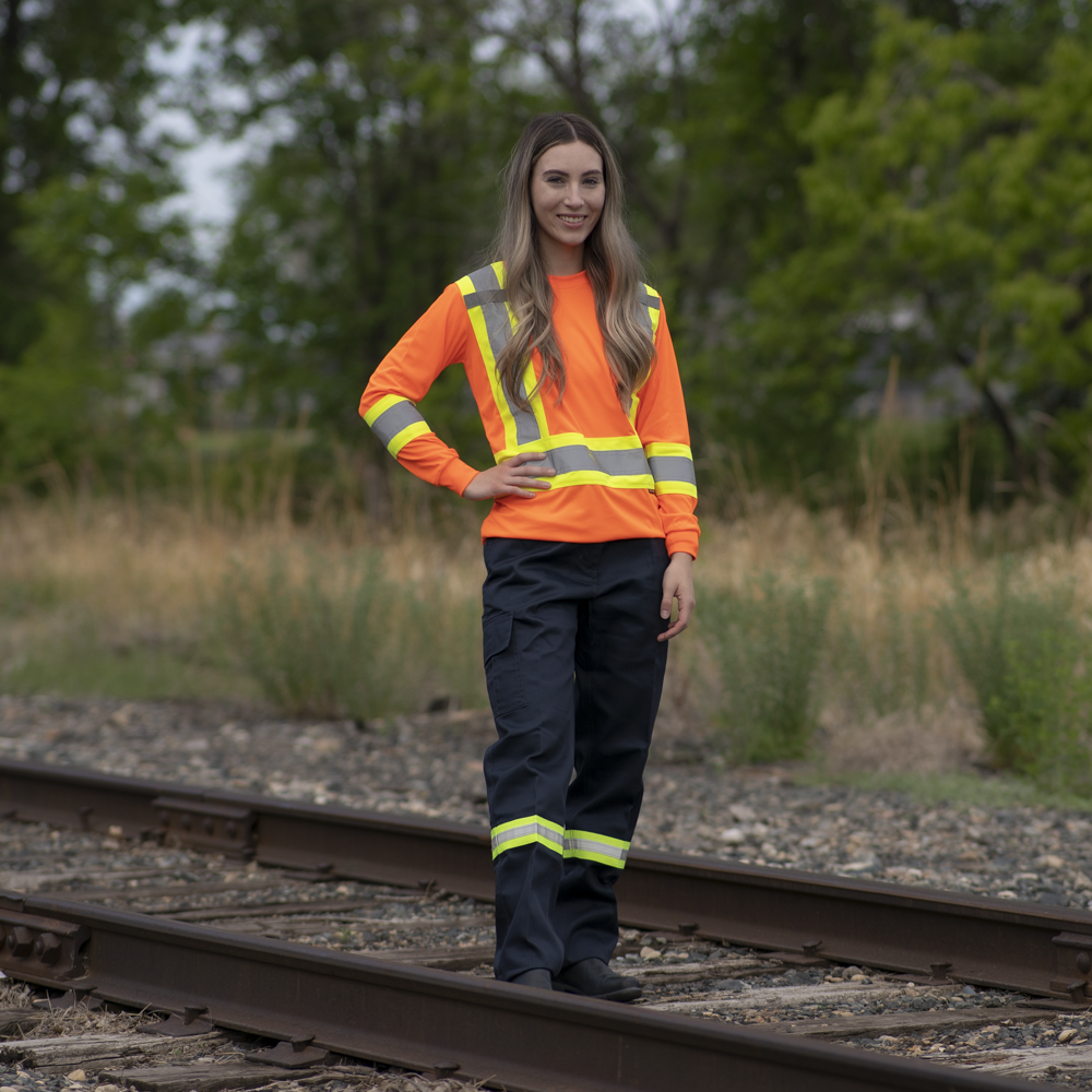 Image of MWG Women's Hi-Vis Long Sleeve T-Shirt. Women's Long Sleeve shirt is bright orange with yellow/silver/yellow reflective tape on torso and sleeves to meet high-visibility standard CSA Z96-15. Model is wearing Hi-Vis Shirt with navy Hi-Vis cargo pants.