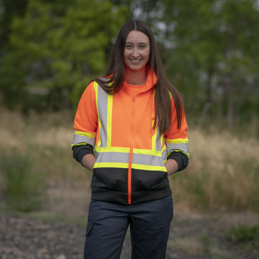 Image of MWG Women's Hi-Vis Hoodie. Women's Hi-Vis Hoodie is bright orange and black in colour with yellow/silver/yellow reflective tape on torso and sleeves to meet high-visibility standard CSA Z96-15. Model is wearing Women's Hoodie with MWG COMFORT WEAVE Women's FR Utility Pant.