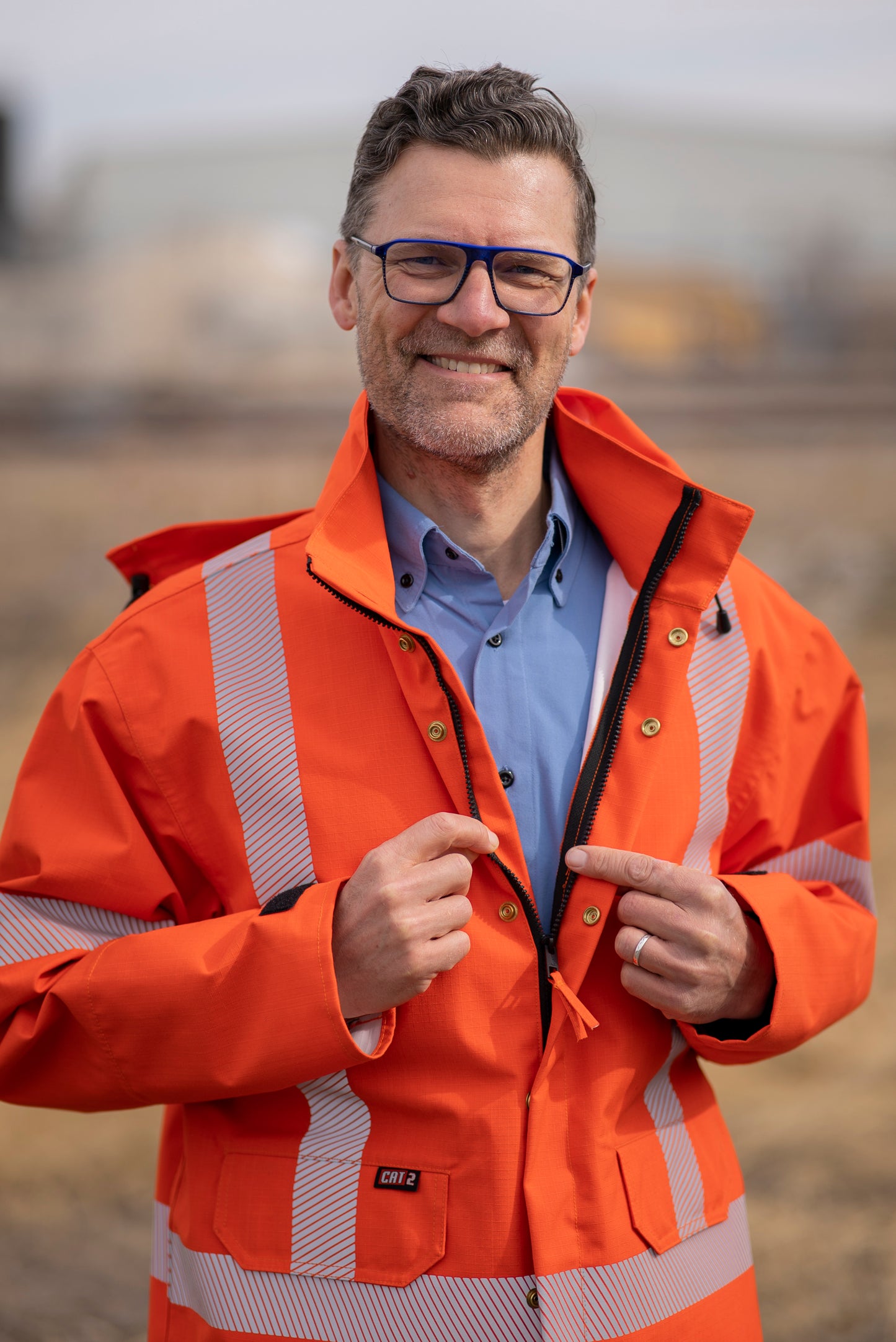 Image of MWG STORMSHIELD FR Rain Proof Jacket. MWG STORMSHIELD FR Rain Proof Jacket is bright orange in colour with silver segmented reflective tape on torso and arms to meet high-visibility standard CSA Z96-15. MWG FR rain jacket has two front pockets with flaps, front zipper with an orange pull, gold buttons to close storm flaps over zipper, and a detachable hood. Model is wearing MWG STORMSHIELD FR Rain Jacket over a bright blue MWG COMFORT WEAVE FR Uniform Shirt.