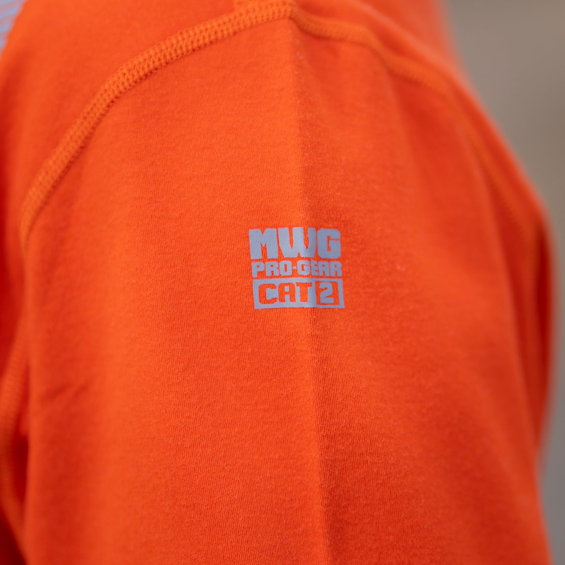 Image of MWG logo and CAT 2 rating on MWG EVOLUTION long-sleeve t-shirt. Logo is silver in colour on bright orange flame-resistant (FR) fabric. MWG EVOLUTION is a lightweight inherent FR fabric.
