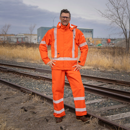 Image of MWG STORMSHIELD FR Rain Proof Jacket. MWG STORMSHIELD FR Rain Proof Jacket is bright orange in colour with silver segmented reflective tape on torso and arms to meet high-visibility standard CSA Z96-15. MWG STORMSHIELD FR Rain Jacket has two front pockets, a detachable hood, and velcro closures on cuffs. Model is wearing MWG STOMSHIELD FR rain jacket with orange MWG STORMSHIELD FR rain proof bib.