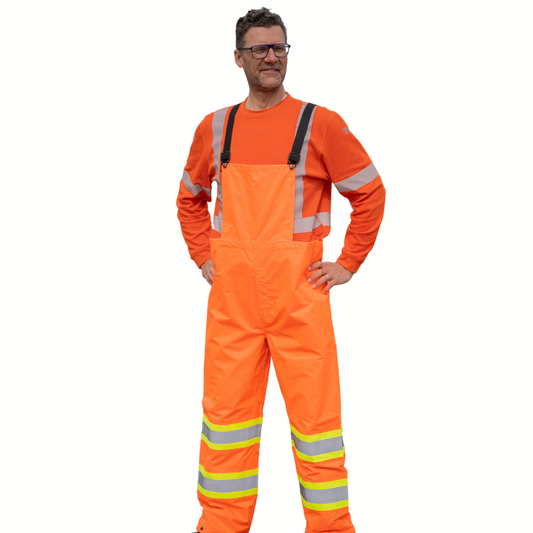 Image of MWG Hi-Vis bib rain pant. Hi-Vis bib rain pant is bright orange in colour with yellow/silver/yellow reflective tape on lower leg to meet high-visibility standard CSA Z96-15. Model is wearing Hi-Vis bib rain pant over orange MWG EVOLUTION long-sleeve t-shirt.
