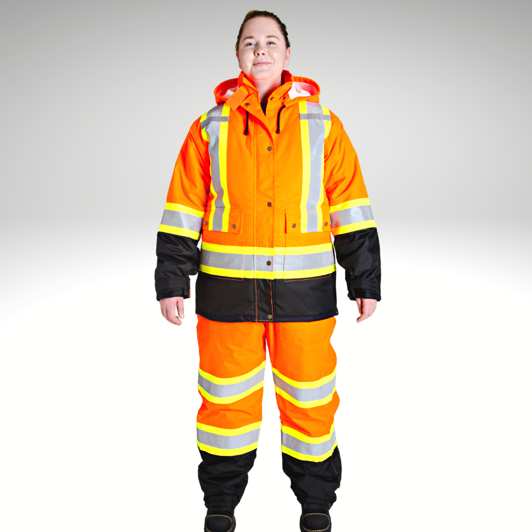 Image of MWG Women's Hi-Vis Insulated Parka Jacket. Women's Hi-Vis Parka is bright orange and black. Yellow/silver/yellow reflective striping on torso, waist, and sleeves to meet high-visibility standard CSA Z96-15. Model is wearing Women's Hi-Vis Parka with hi-viz insulated bib overalls.