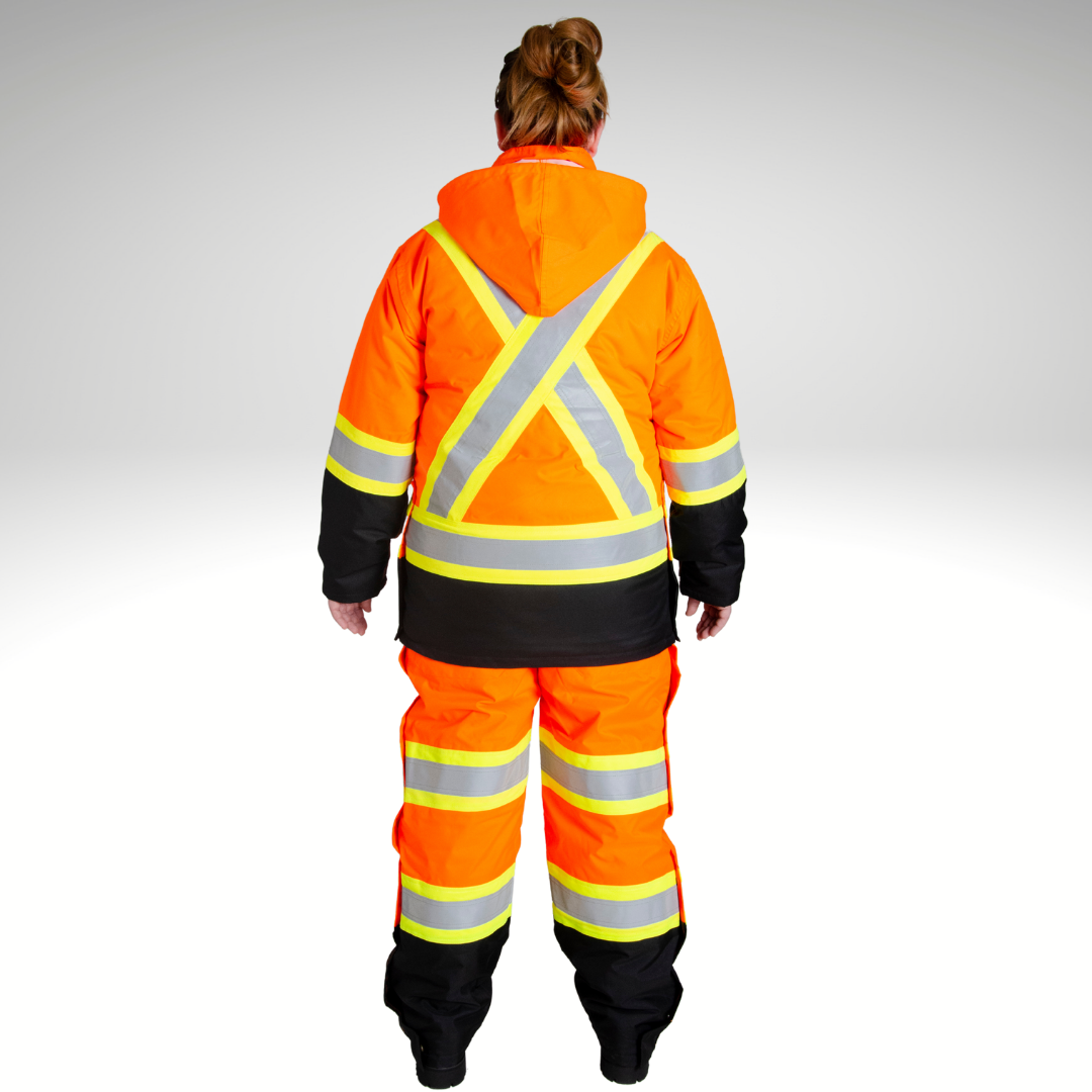 Image of MWG Women's Hi-Vis Insulated Parka Jacket. Women's Hi-Vis Parka is bright orange and black. Yellow/silver/yellow reflective striping on upper back in an X pattern, waist, and sleeves to meet high-visibility standard CSA Z96-15. Model is wearing Women's Hi-Vis Parka with hi-viz insulated bib overalls.