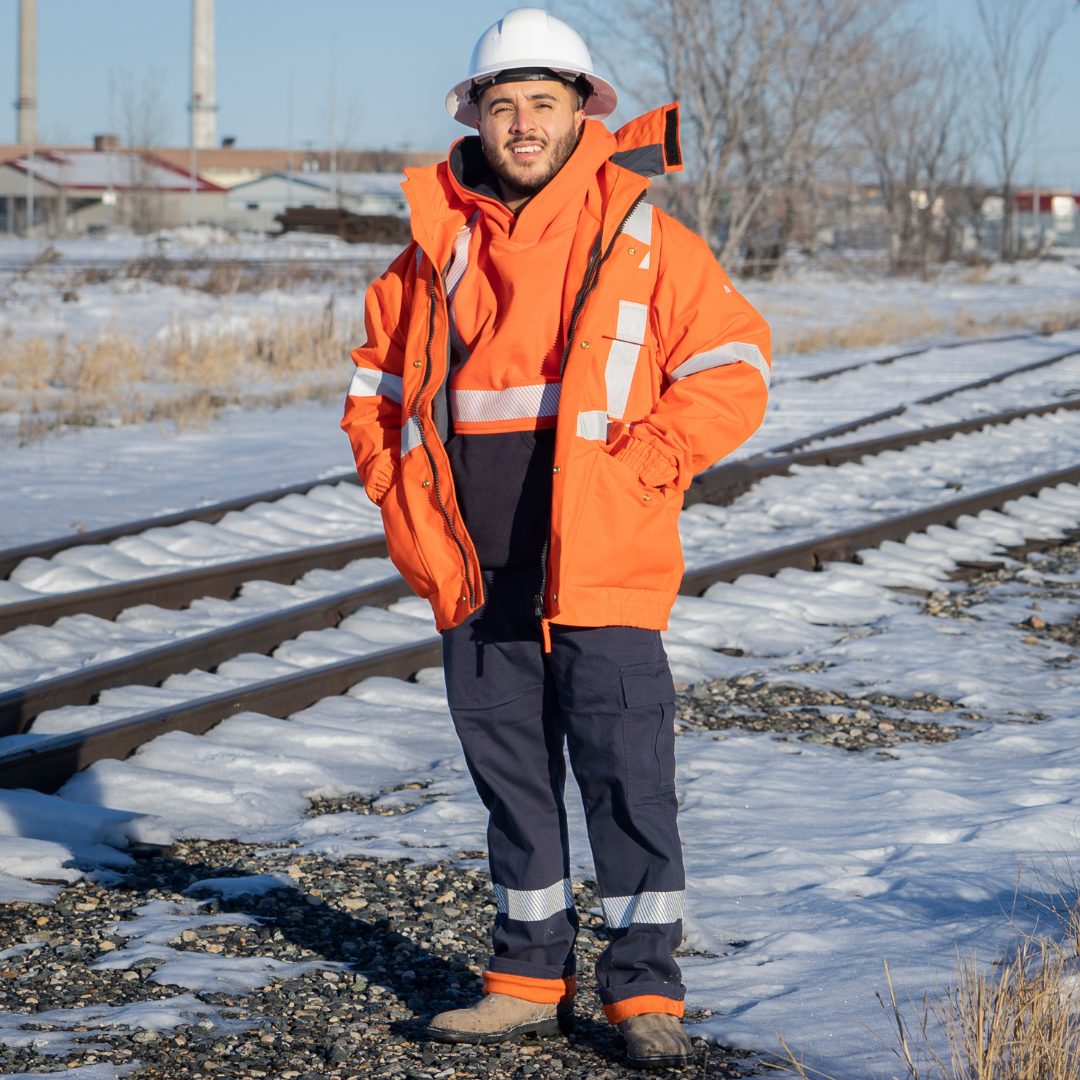 Image of MWG STORMSHIELD FR/AR Winter Jacket. FR Jacket is bright orange with silver reflective striping on torso and sleeves for high-visibility. MWG STORMSHIELD is an inherent flame-resistance ripstop fabric. Model is wearing FR Jacket with navy flame-resistant pants. Men's FR Freezer Jacket zips in for additional warmth.